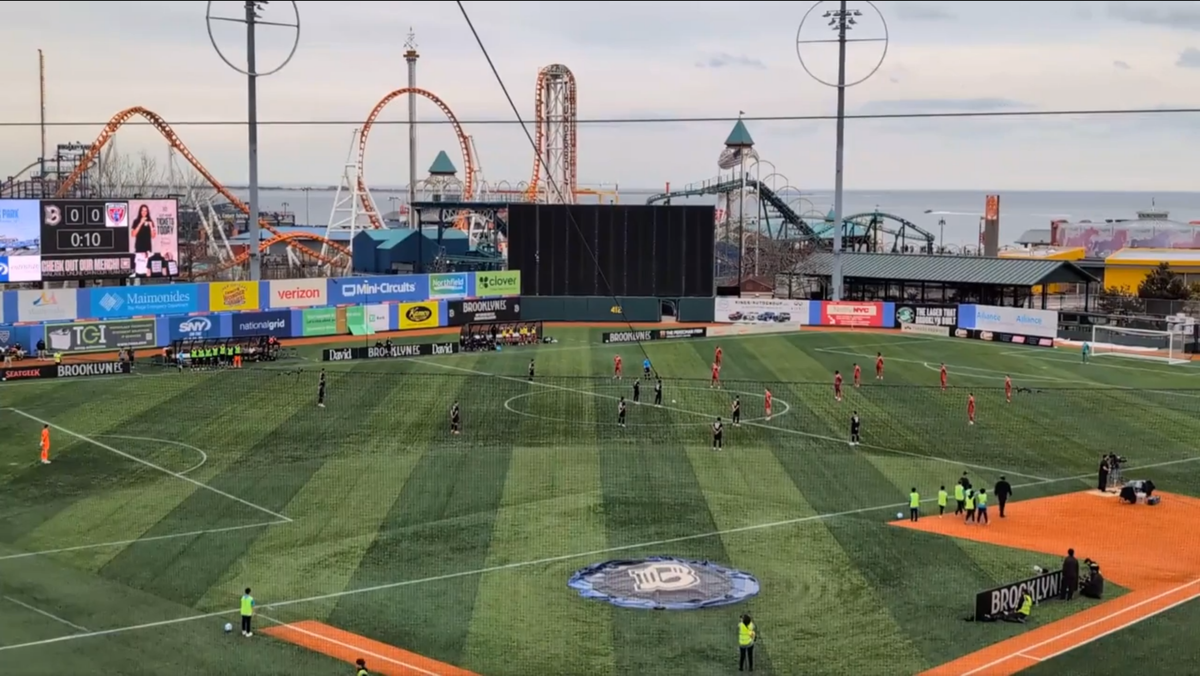 Players for Brooklyn FC and Indy Eleven stand spread out across a soccer field at the opening whistle, none moving.